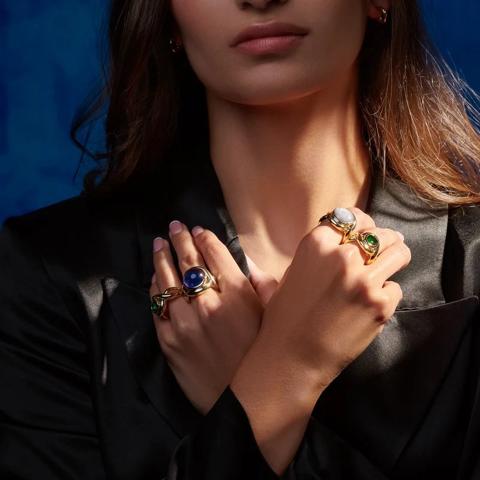 Hands crossed over a black jacket, wearing gold rings set with coloured stones