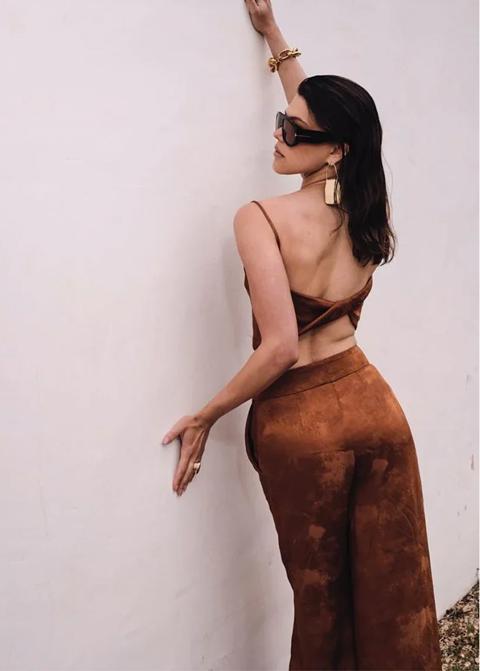 Woman in rust satin trousers and sunglasses leaning on a white wall